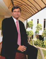 Falgunbhai Patel standing in a covered walkway beside a garden courtyard, wearing a dark suit.